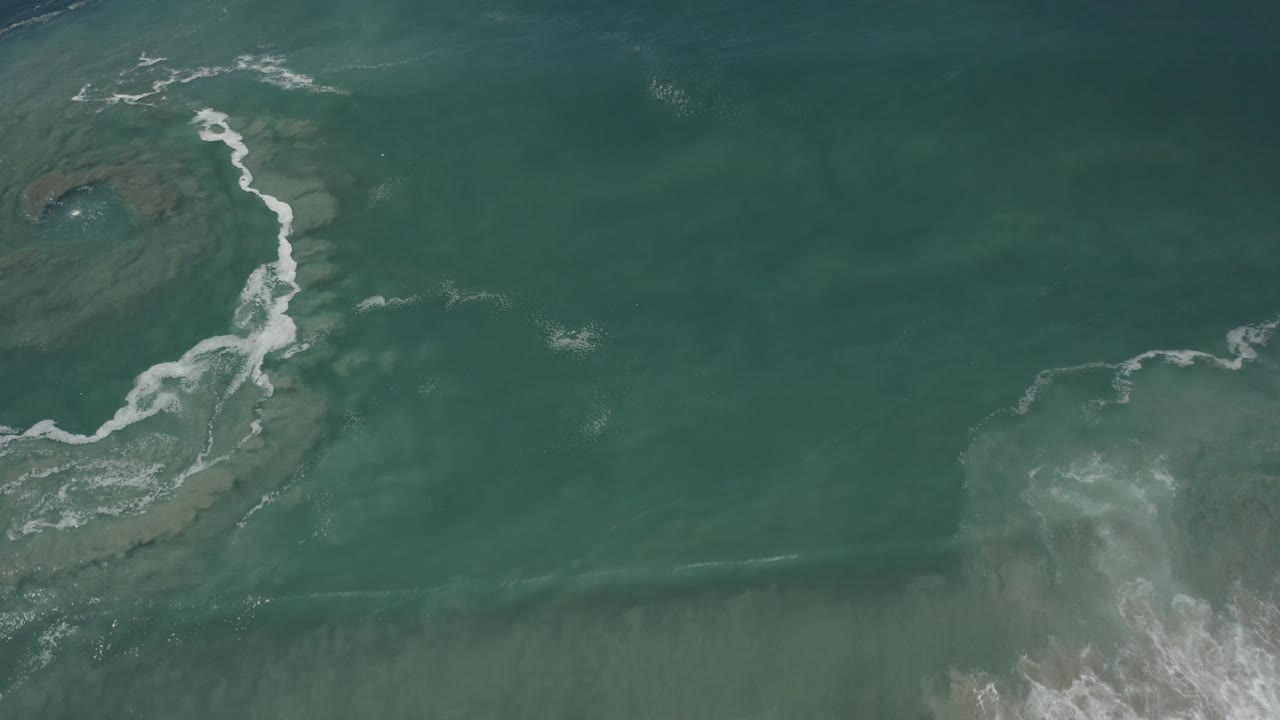 Top-down surf foam wraps around a dark reef shape in green ocean water.