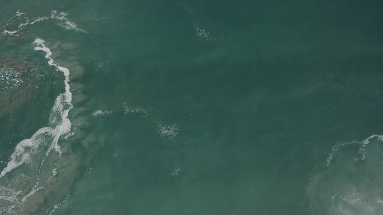 Overhead view of green-blue ocean water in San Jose del Cabo, Mexico, with white sea foam entering from the left side and no visible people.