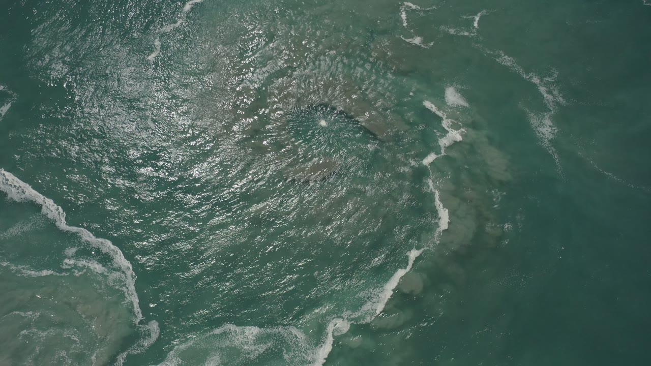 Top-down view of green ocean water in San Jose Del Cabo, Mexico, with diagonal white foam streaks, bright surface glare, and no visible people.
