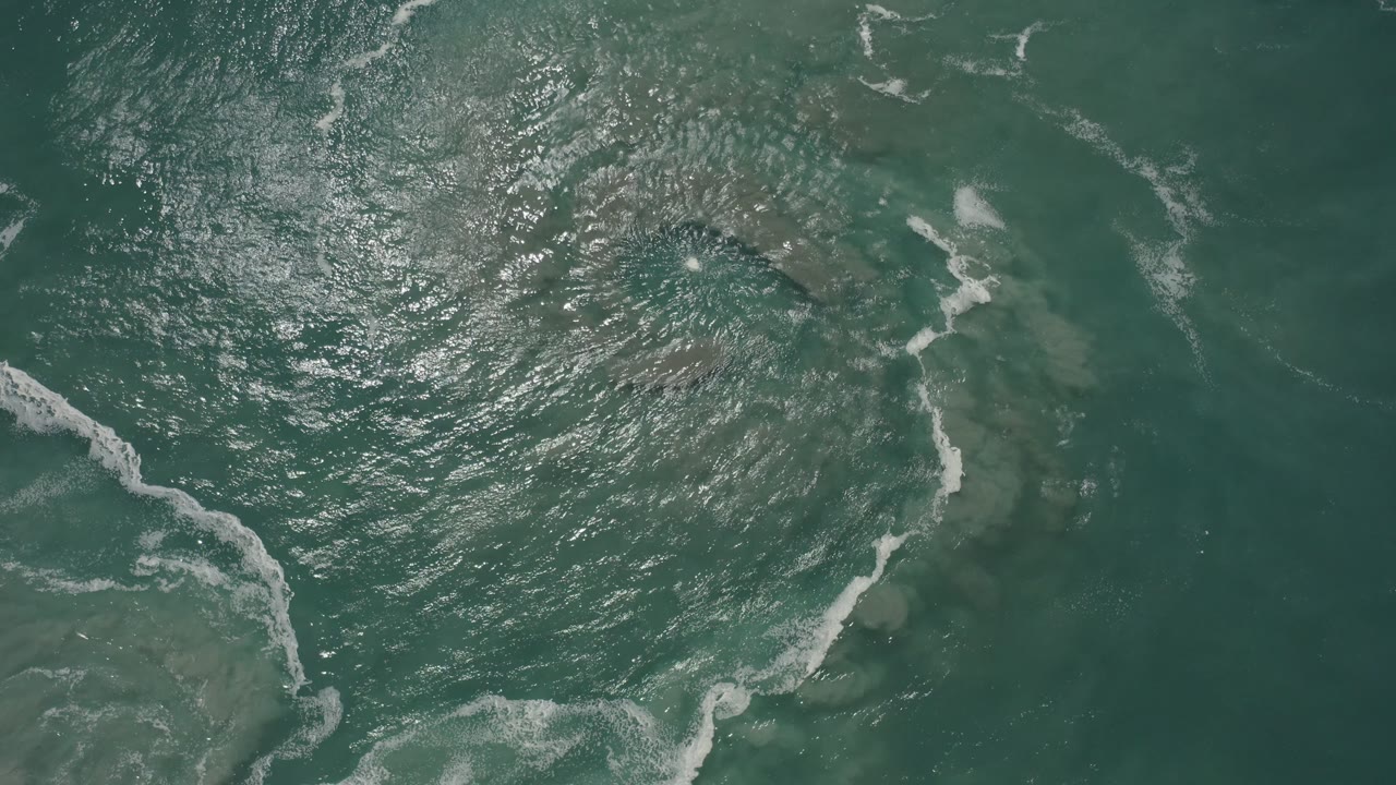 Overhead green sea surface in San Jose Del Cabo, Mexico, with broken foam lines spread farther across the left side and no visible people.