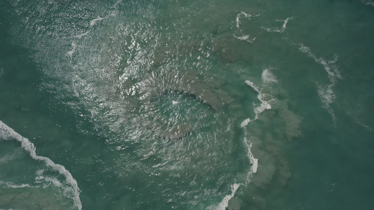 Top-down drone view over green-blue ocean near San Jose del Cabo, Mexico, with bright sun reflections, submerged shapes, thin white foam, and no visible people.