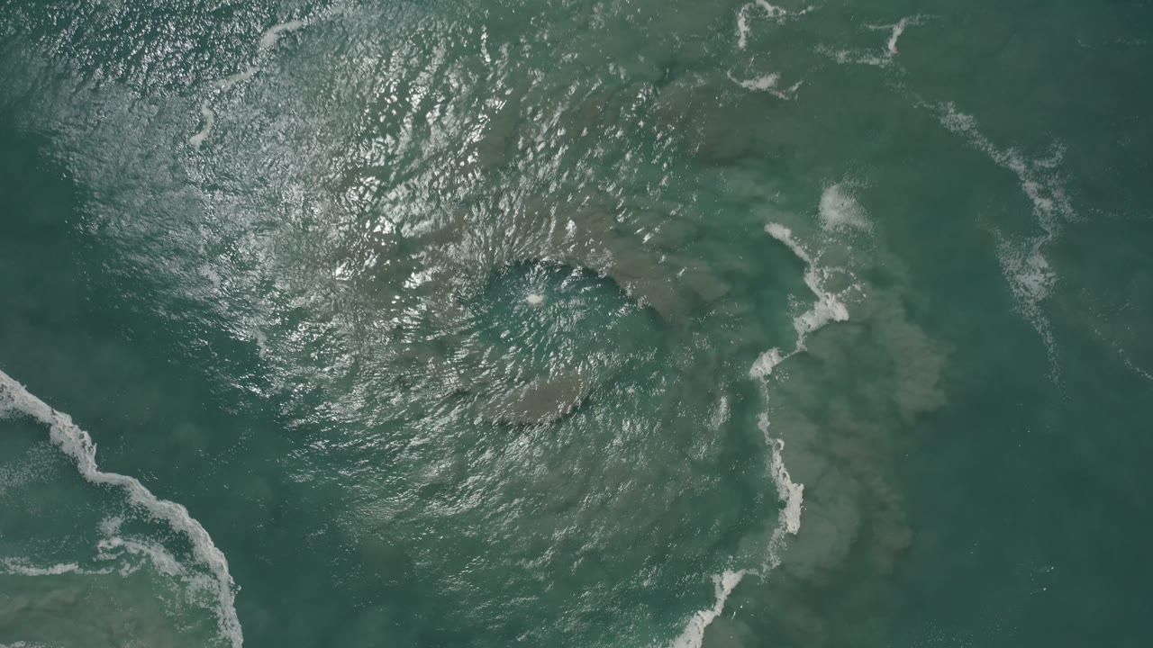 Sunlit ocean viewed from above with diagonal surf foam and cloudy underwater color.