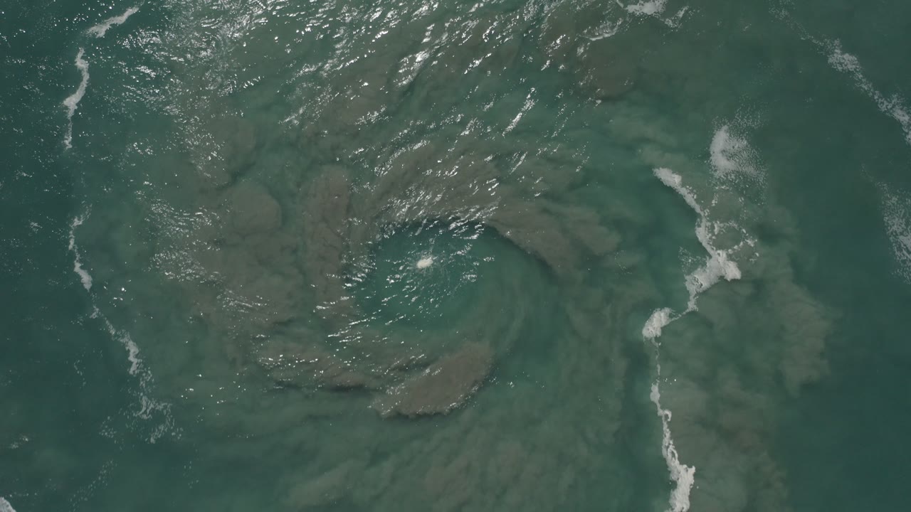 Overhead view in San Jose del Cabo, Mexico, showing green water over a circular sand or mud formation with white ripples near the top and no visible people.