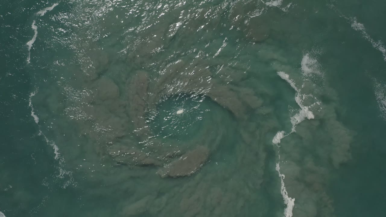 A slightly shifted overhead frame in San Jose del Cabo, Mexico, keeps the circular sand or mud shape under green water while bright surface ripples gather across the upper left, with no visible people.