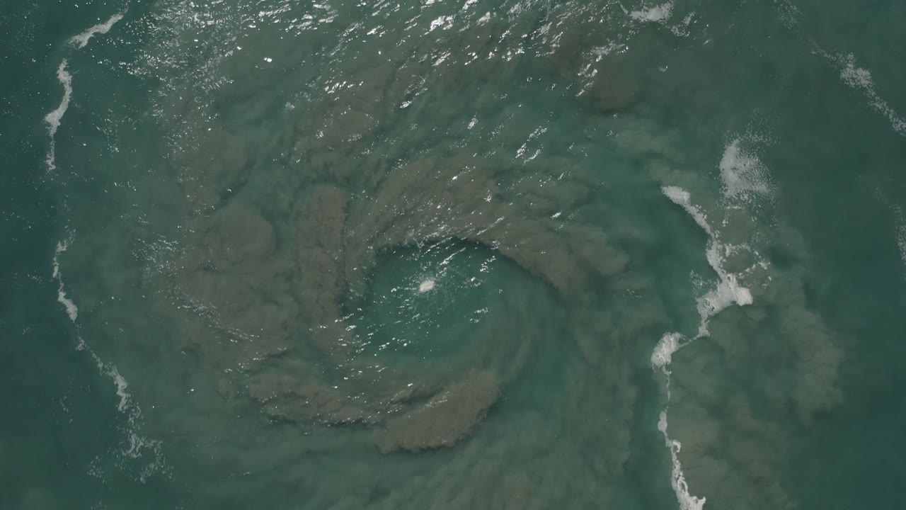 Green coastal water over a rounded sand or mud swirl with white foam streaks.