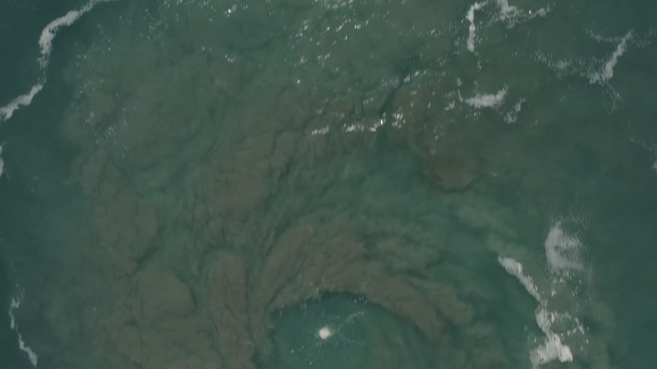 Overhead view of emerald green ocean water in San Jose del Cabo, Mexico, with pale seabed shapes below the surface, scattered white foam, and no visible people.