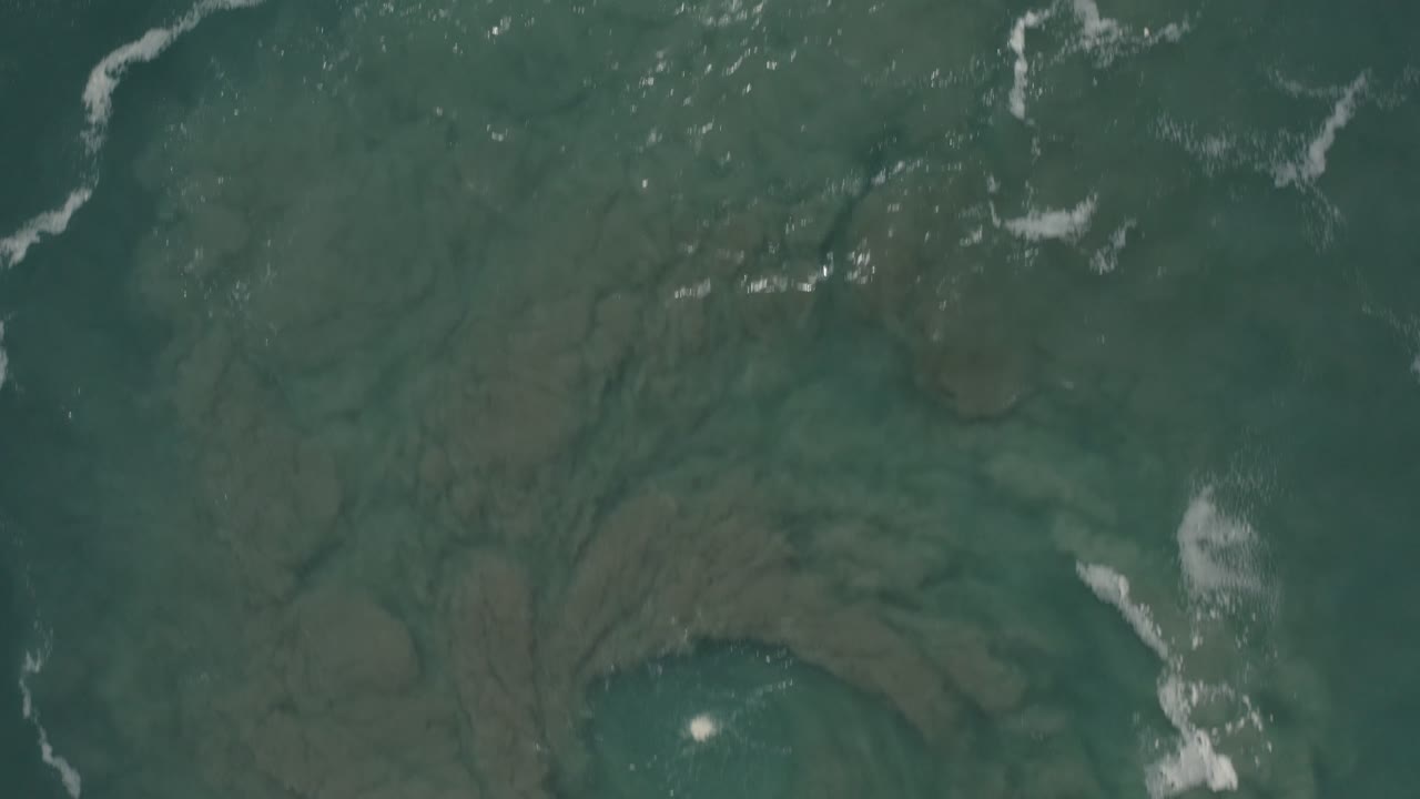 Green ocean surface in San Jose del Cabo, Mexico, seen from above with muted seabed patterns, narrow foam streaks, and no visible people.
