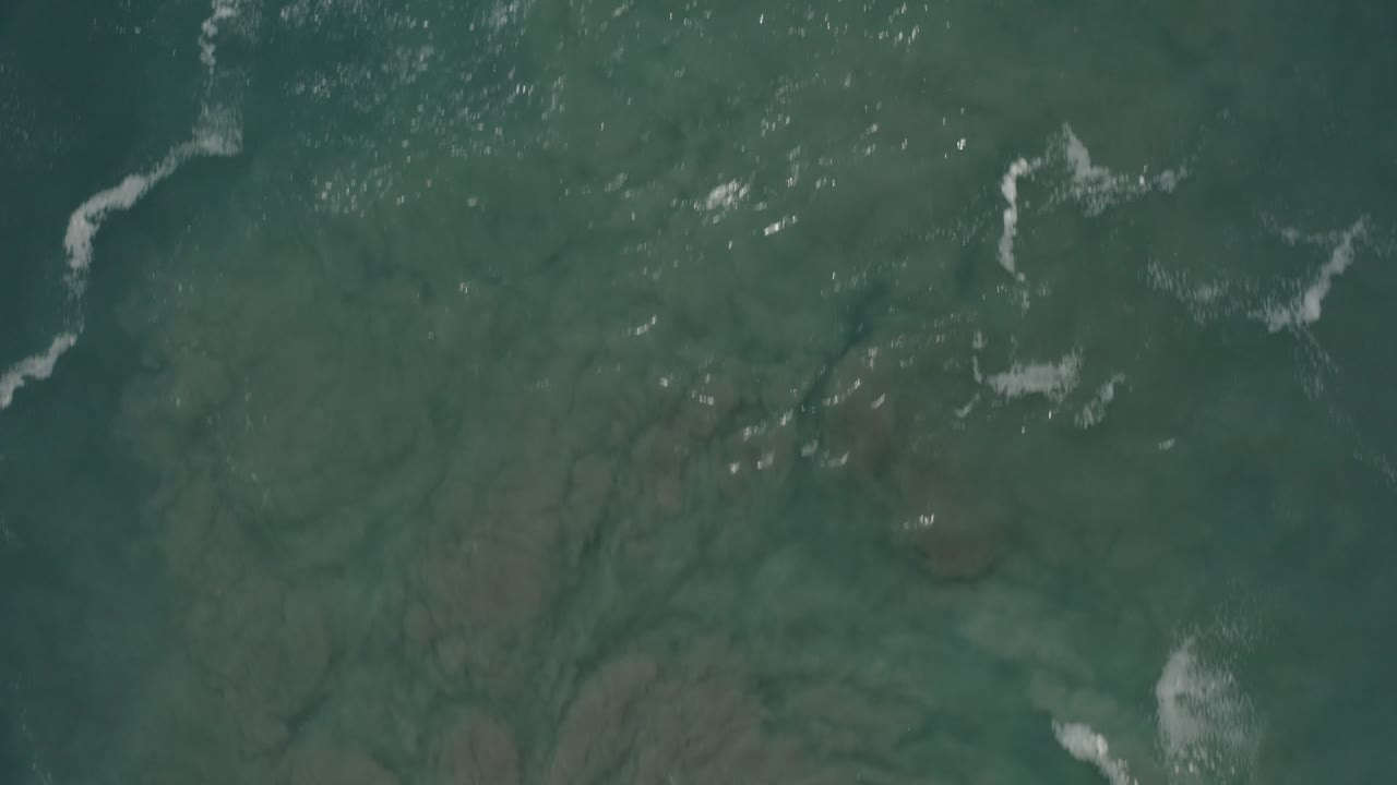 Aerial view of San Jose del Cabo ocean water with darker seabed patches, thin white foam edges near the top and right side, and no visible people.