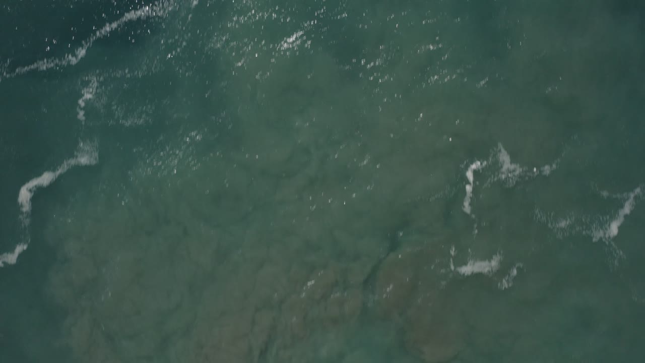 Close overhead view of emerald water in San Jose del Cabo, Mexico, with a broad muted seabed area, scattered foam marks, and no visible people.