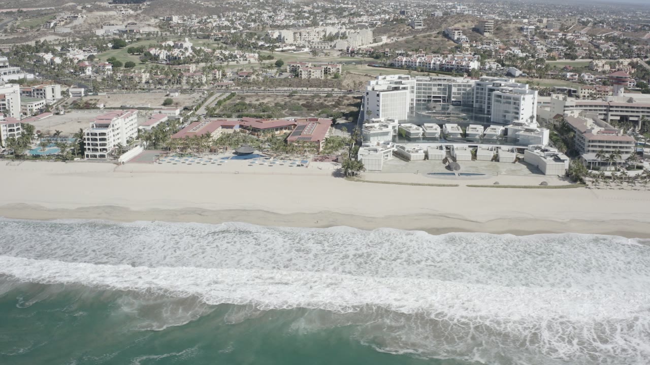 Aerial frame over San Jose del Cabo, Mexico, with teal surf in the foreground, pale sand, white beachfront hotels, inland development, and no visible people.