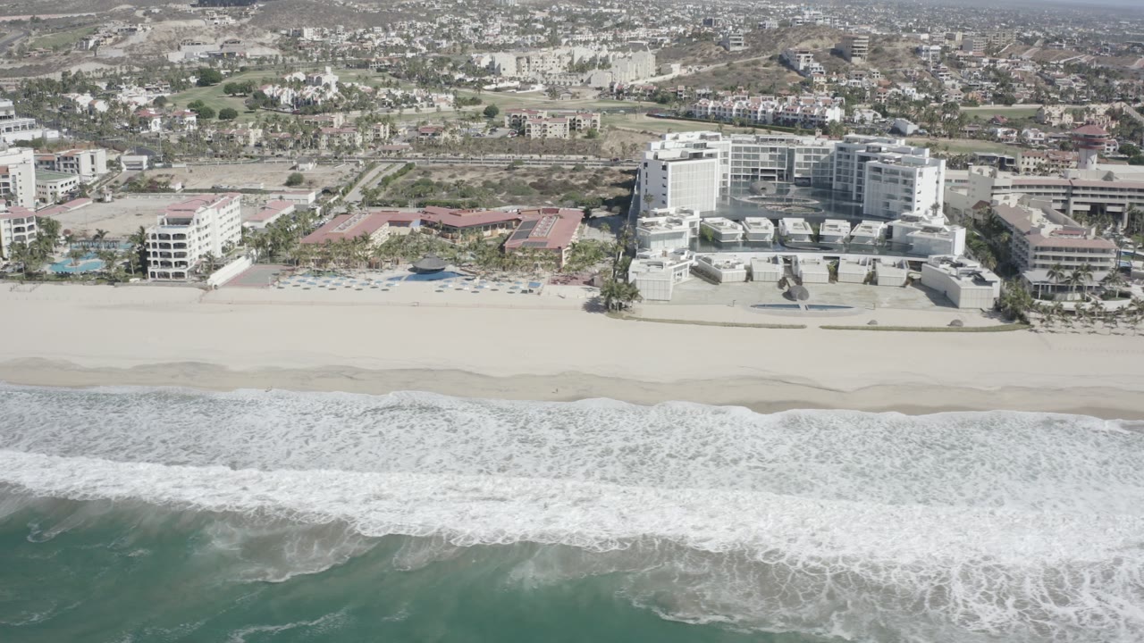 San Jose del Cabo aerial view with foamy waves spread across the lower frame, a broad empty-looking beach, resort buildings near center, and no visible people.