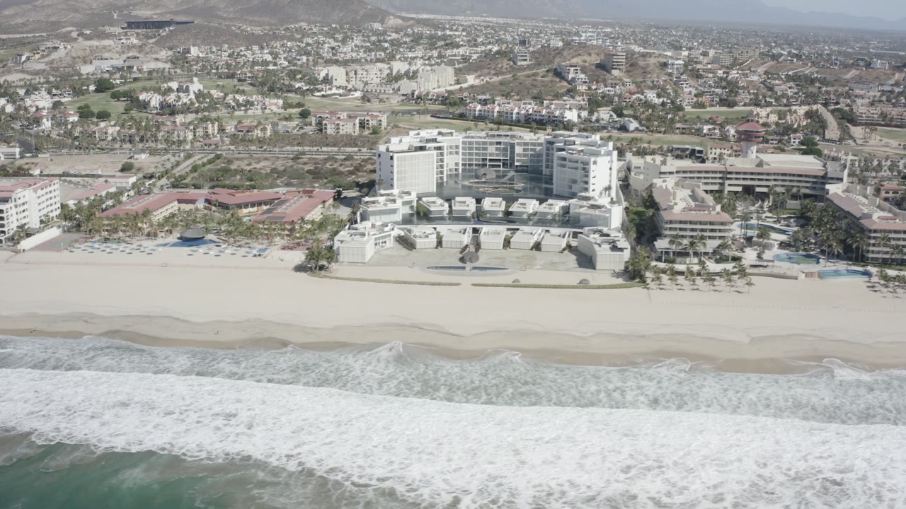 A wide aerial frame shows white beachfront buildings along a sandy beach in San Jose del Cabo, Mexico, with foamy surf in front and inland hills behind, with no visible people.