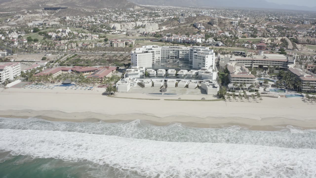 The aerial view centers the white beachside complex in San Jose del Cabo, Mexico, while a band of breaking waves fills the lower edge and dry hills sit beyond the town, with no visible people.