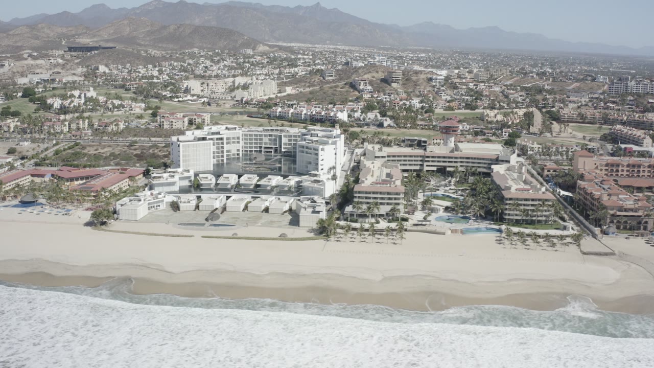 Aerial view of beachfront buildings, surf, city blocks, and distant mountains.