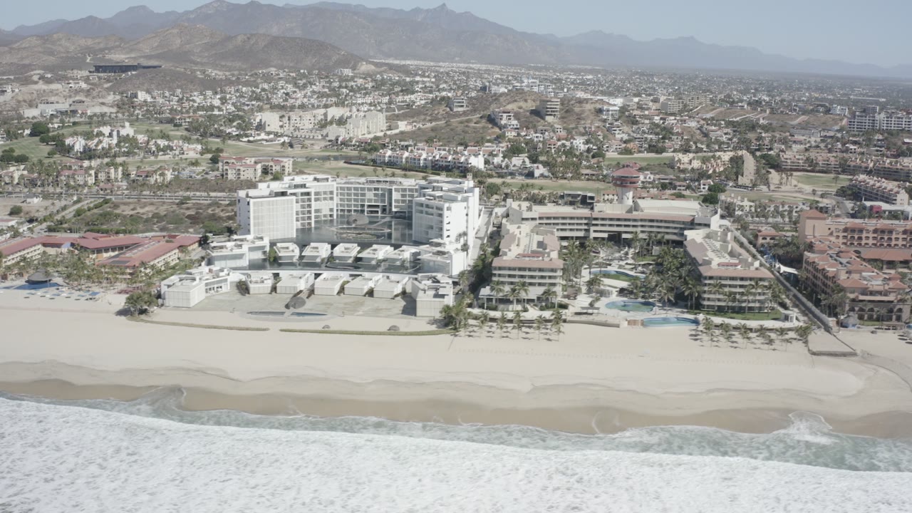 Aerial frame in San Jose del Cabo, Mexico, showing a large white beachfront building, neighboring low-rise structures, surf at the bottom edge, hazy mountains beyond the city, and no visible people.