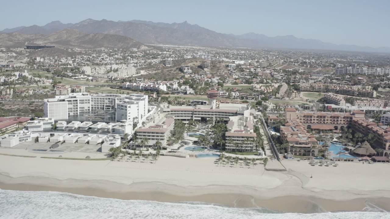 Aerial view of beach, surf, resort buildings, town, and mountains.
