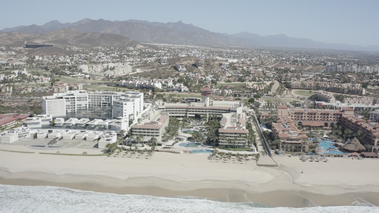 High oblique view in San Jose Del Cabo, Mexico, showing white beachfront buildings, terracotta resort roofs, a strip of sand, breaking waves, inland streets, and no visible people.