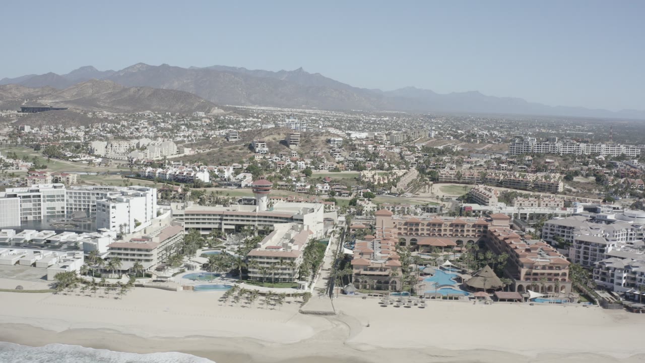 Wide aerial frame over San Jose del Cabo with sandy beach at the bottom, resort buildings and pools in the middle distance, hazy mountains beyond, and no visible people.