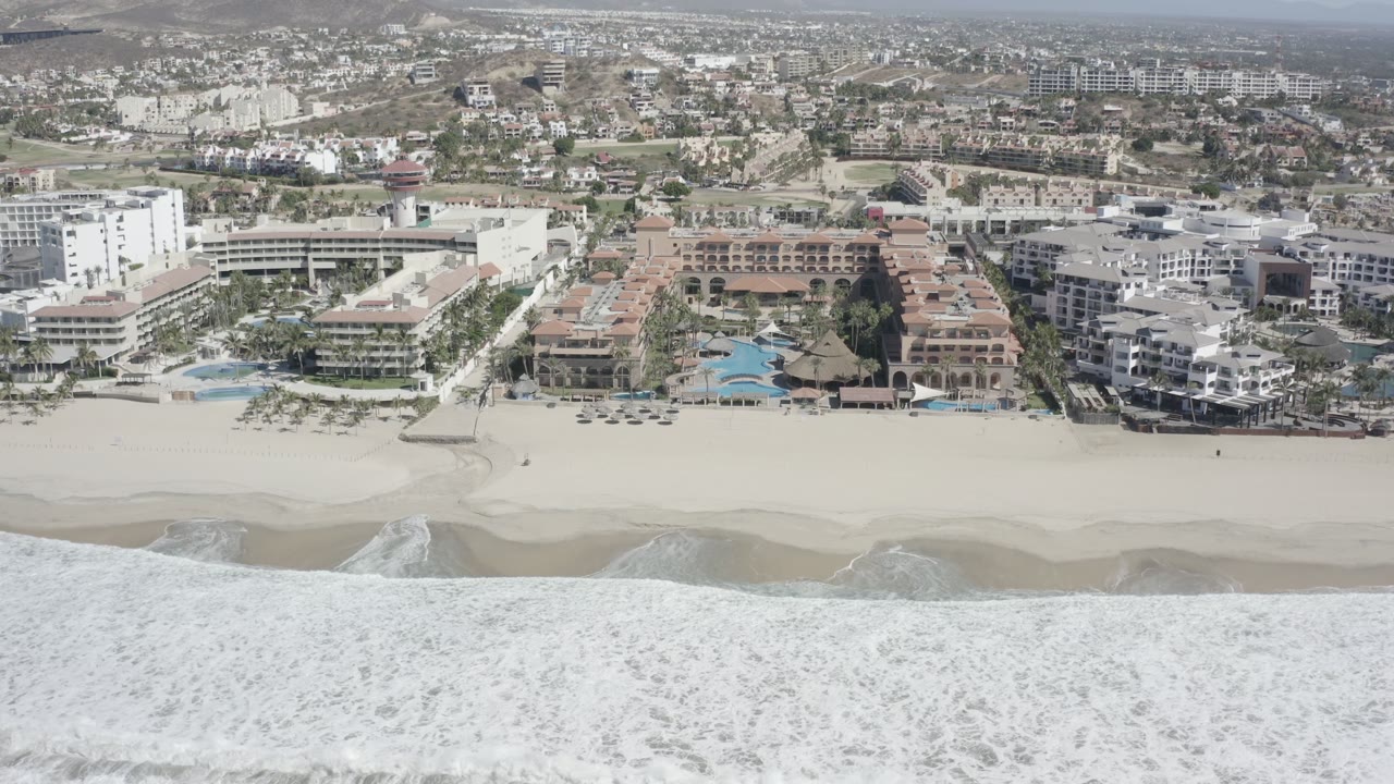 Opening aerial frame over San Jose Del Cabo surf and sand, with beachfront apartments, pools, palms, inland streets, dry hills, and no visible people.