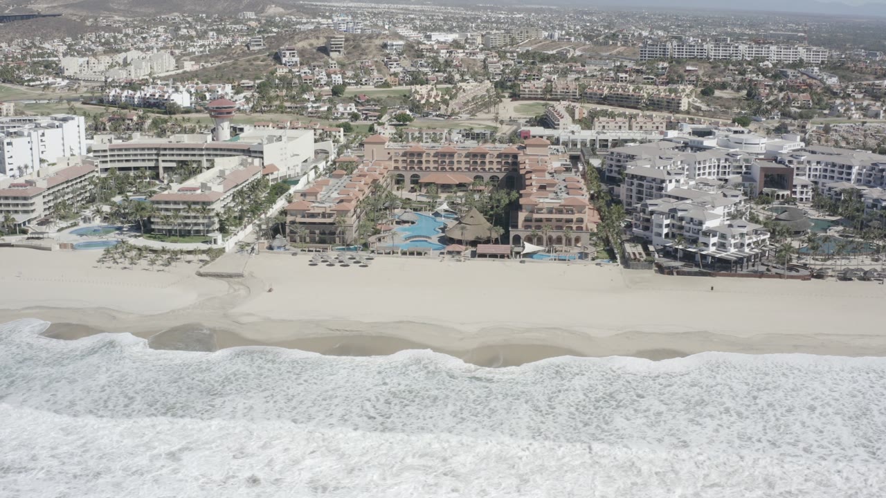 The next aerial frame shows foamy water lower in the image, smooth sand through the center, and clustered beachfront buildings and pools in San Jose del Cabo, with no visible people.