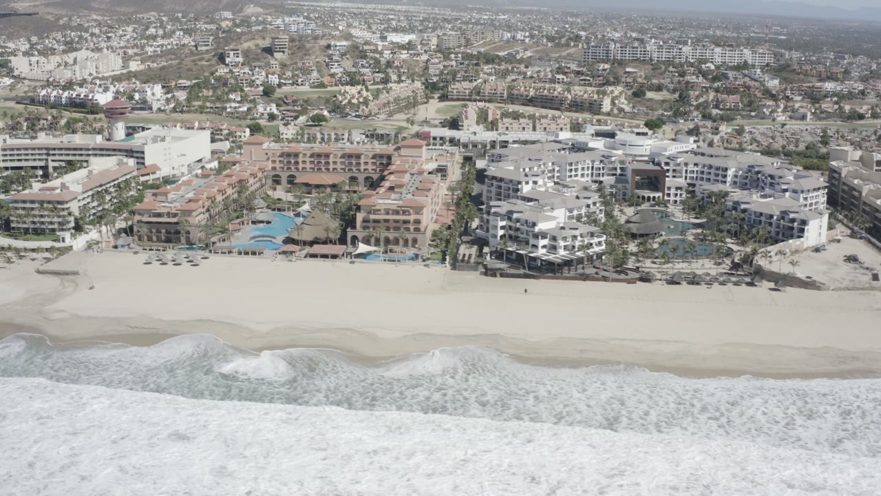 Aerial frame over San Jose Del Cabo showing white surf in the foreground, a wide pale beach, and beachfront resort buildings with no visible people.
