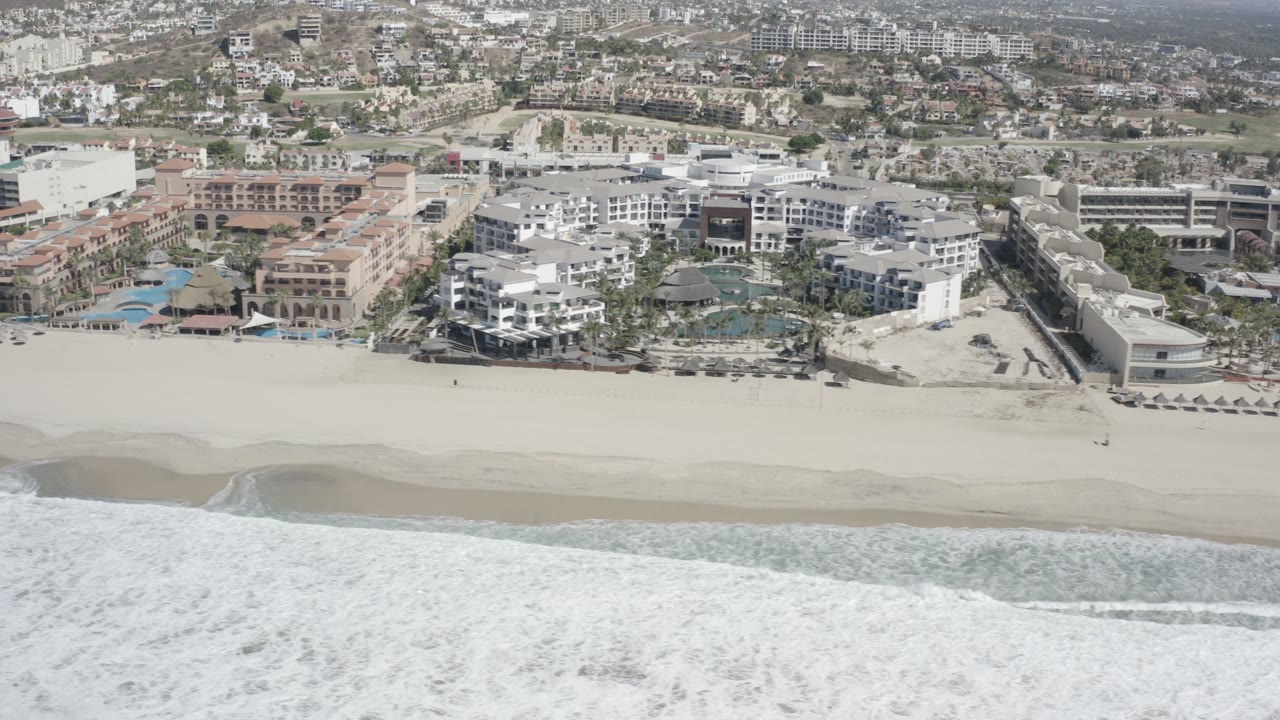 Aerial frame in San Jose del Cabo, Mexico, showing foamy waves closest to camera, a wide empty-looking beach, clustered resort structures, blue pools, and no visible people.