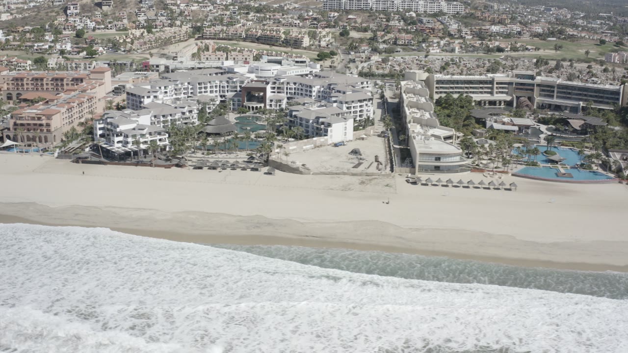 Aerial frame from San Jose Del Cabo showing foamy surf below a wide sandy beach, resort buildings, turquoise pools, and no visible people.