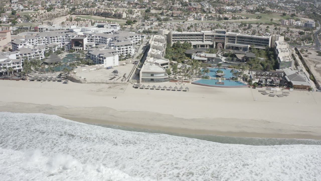 High aerial frame over San Jose Del Cabo showing breaking white waves below a broad sandy beach and a resort complex with blue pools and low buildings behind it.