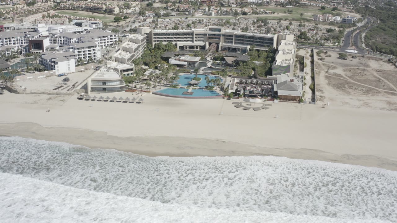 High aerial frame over San Jose del Cabo, Mexico showing foamy surf below, a wide white sand beach, resort-style buildings, pools, roads, and no visible people.