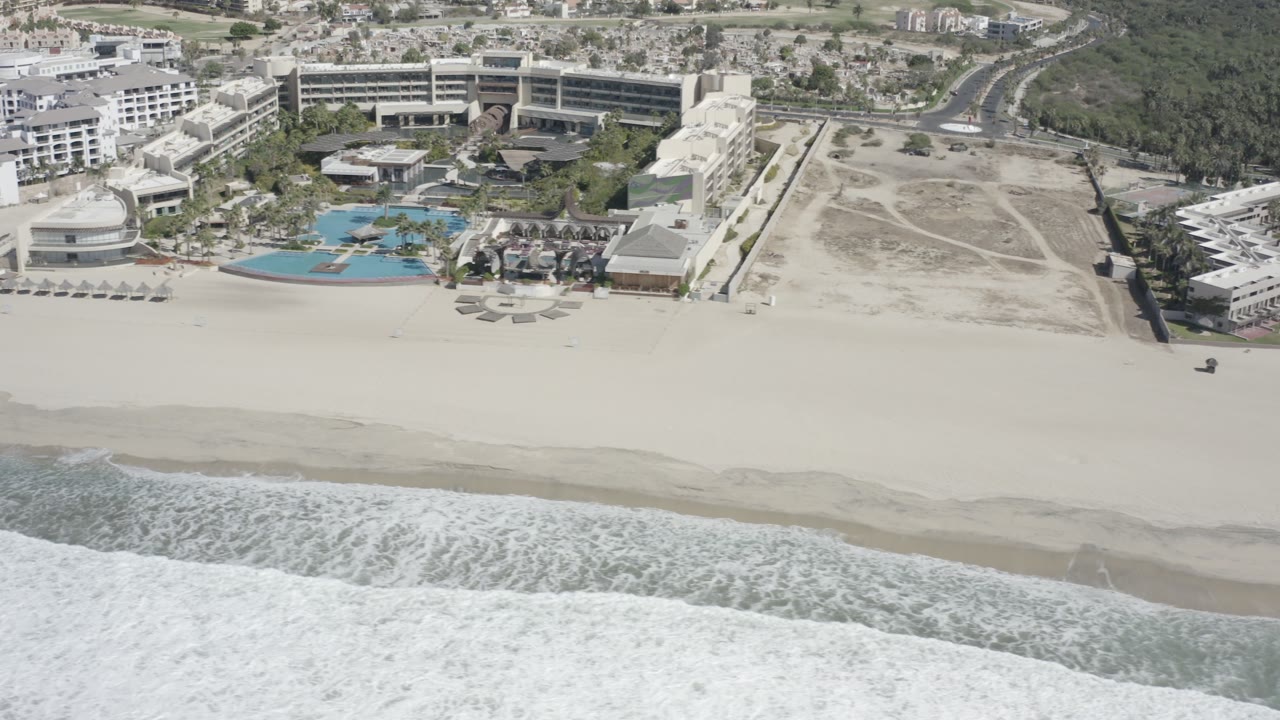 Aerial frame over San Jose Del Cabo showing foamy surf at the bottom, a wide pale beach, blue resort pools, large hotel buildings, vacant sandy lots, and no visible people.