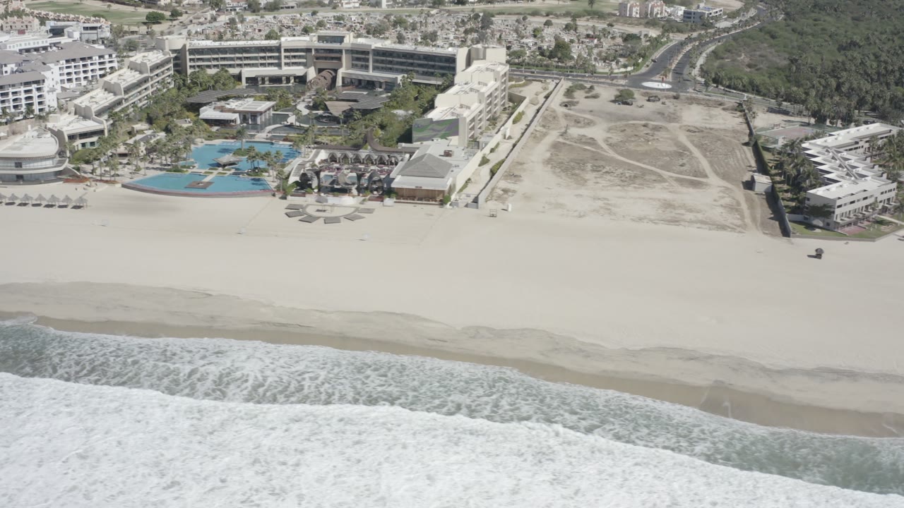Beachfront resort area with waves, pools, hotel buildings, and open lots.