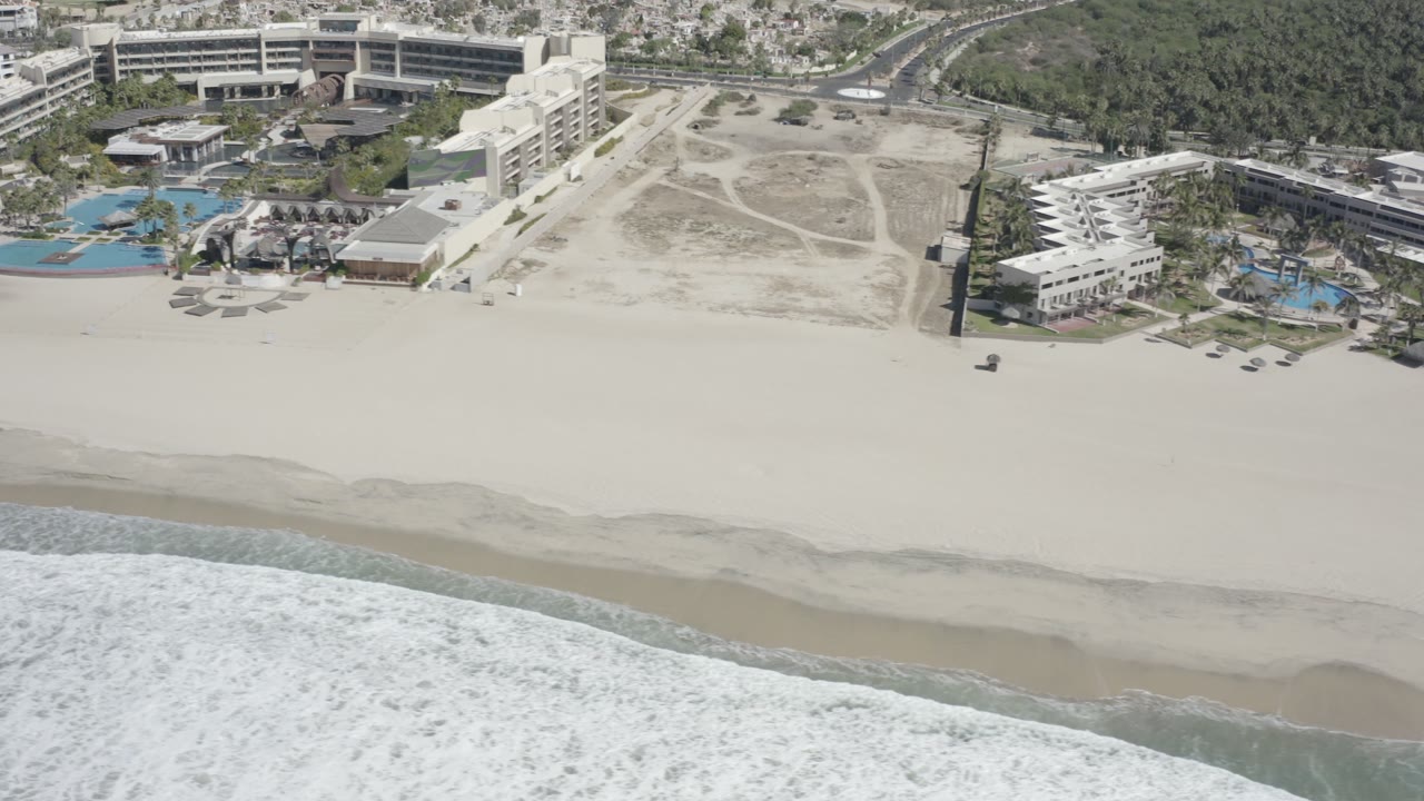 Aerial frame of a San Jose del Cabo beach with white surf at the bottom, wide pale sand in the center, resort buildings and pools beyond, and no visible people.