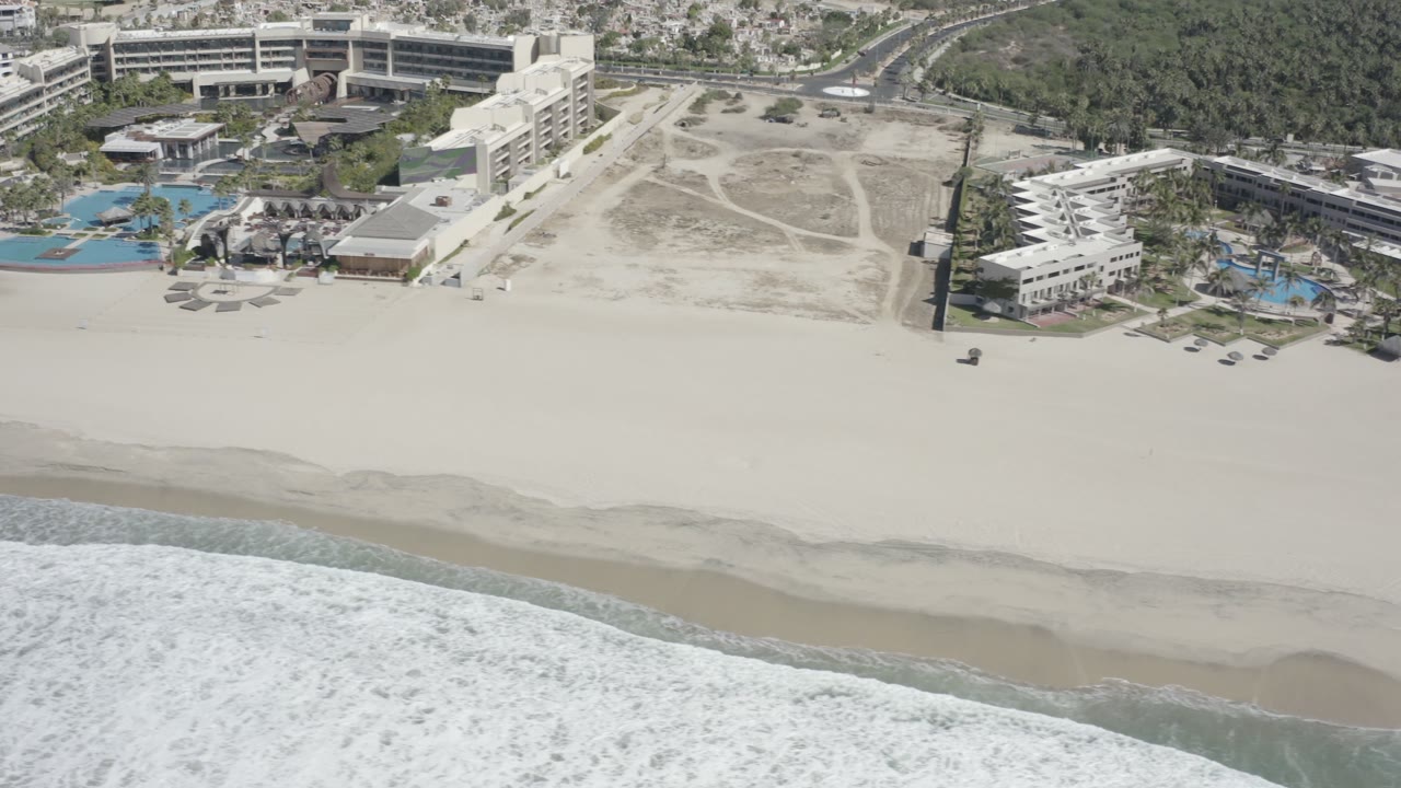 Aerial view over San Jose del Cabo shoreline as foamy waves reach the lower edge, with broad empty sand and resort buildings set back from the beach, no visible people.