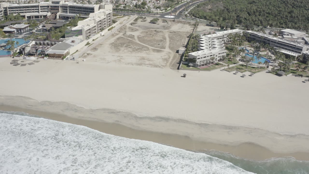Drone-style view of a sandy beach, rolling surf, and coastal buildings.