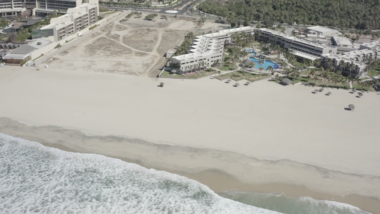 Aerial frame from San Jose del Cabo showing foamy surf, pale sand, beachfront buildings, palm-lined grounds, blue pools, and no visible people.