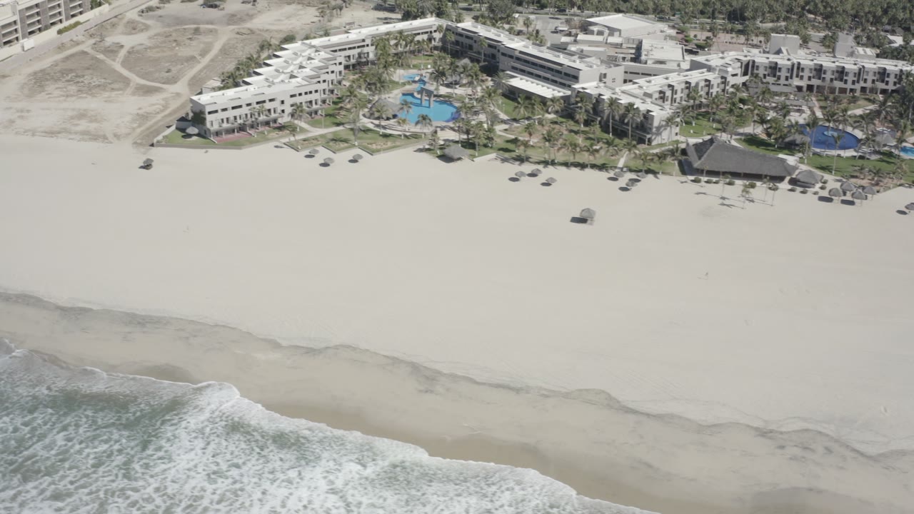 San Jose del Cabo aerial view with white water reaching the lower edge, open sand in the center, and modern beachfront buildings and blue pools along the top, with no recognizable people.