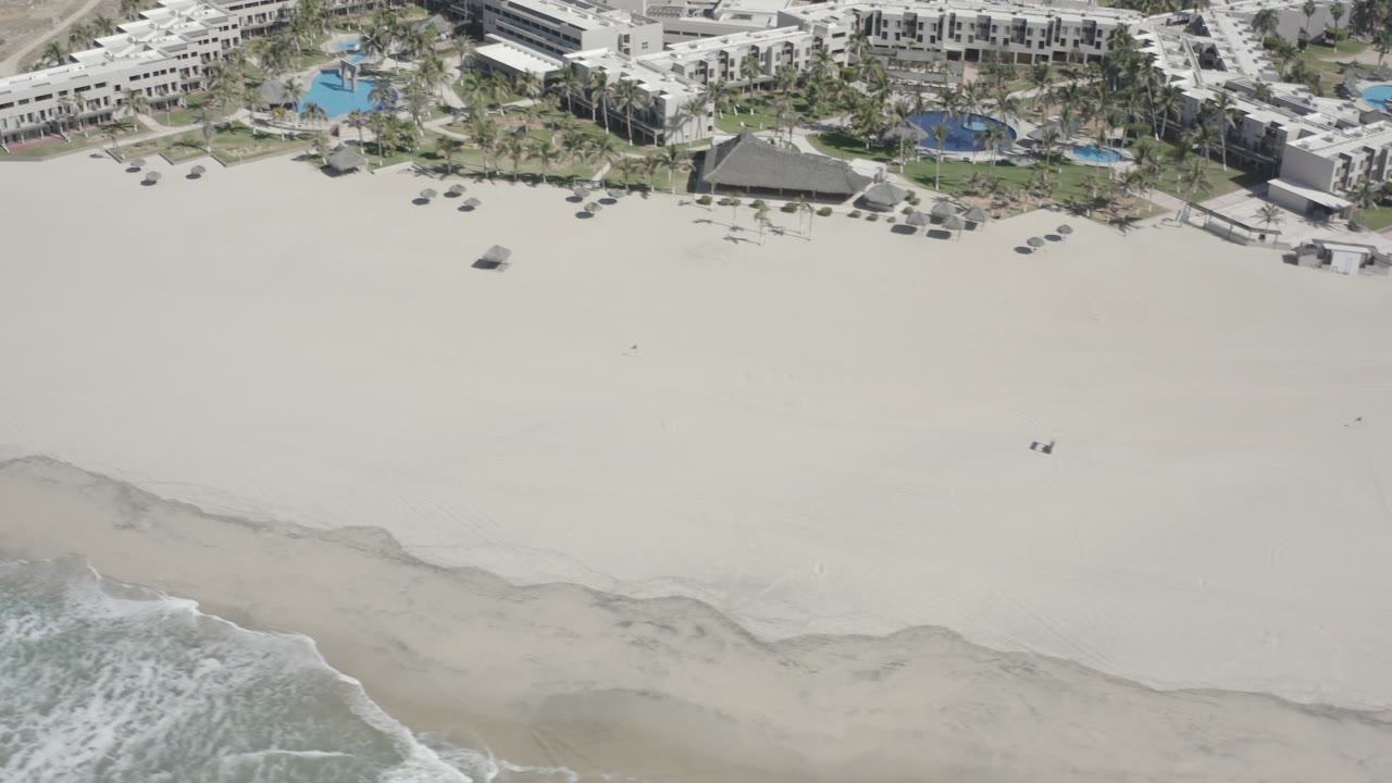 Aerial frame over San Jose del Cabo showing a wide sand field, two small people near the center, beachfront buildings and pools at the top, and waves entering from the bottom left.