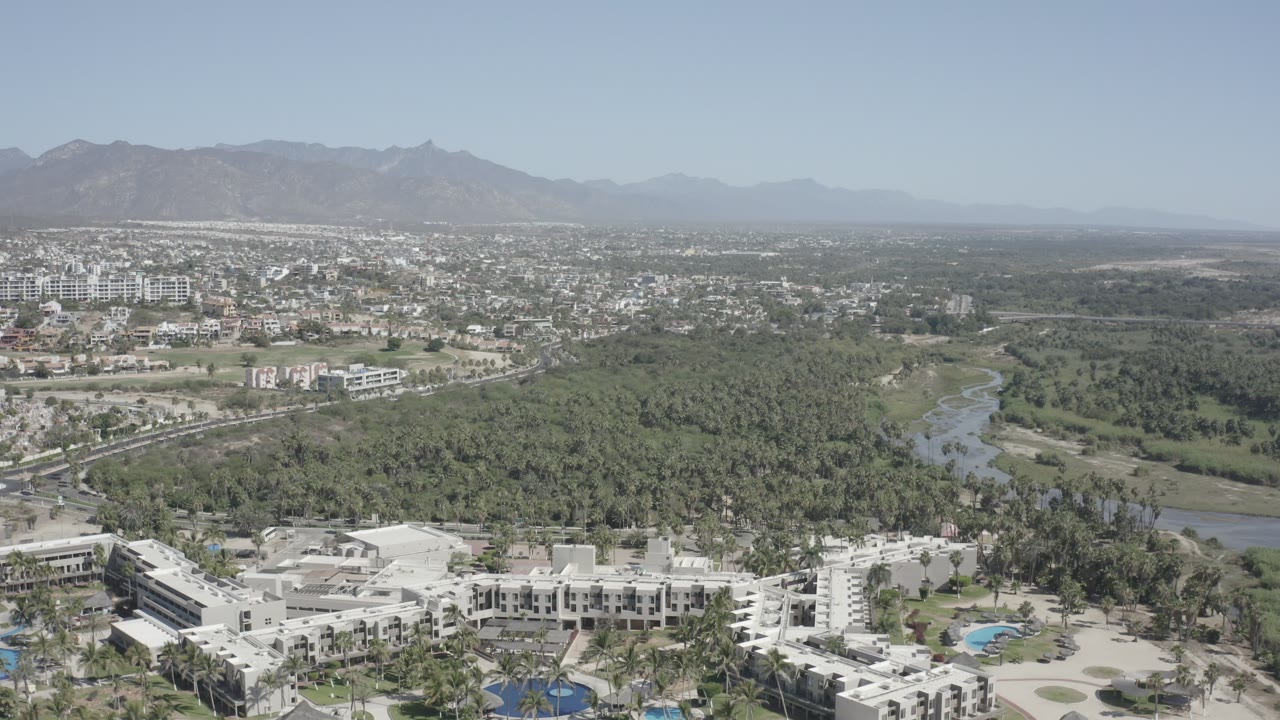 Second aerial frame in San Jose del Cabo showing the park and waterway centered between foreground buildings and the spread of city blocks under a pale blue sky, with no visible people.