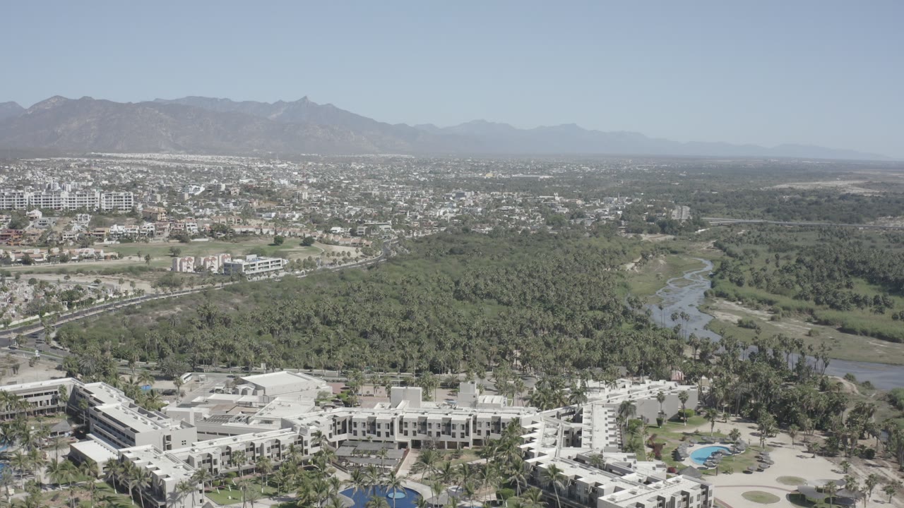 Second aerial frame of San Jose del Cabo with resort-like white buildings at the bottom edge, green vegetation across the center, a narrow water channel, distant development, mountains, and no visible people.