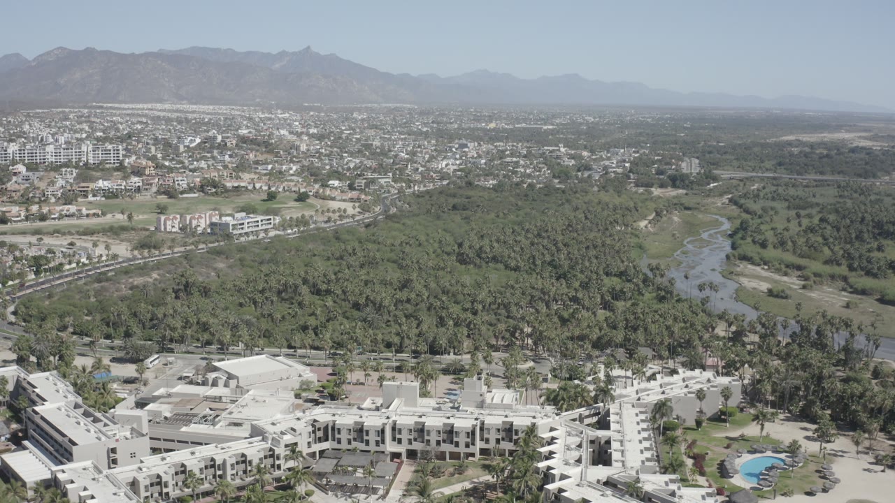 Opening aerial frame over San Jose del Cabo with white low-rise buildings at the bottom, a green palm corridor and winding waterway through the center, city blocks beyond, distant mountains, and no visible people.