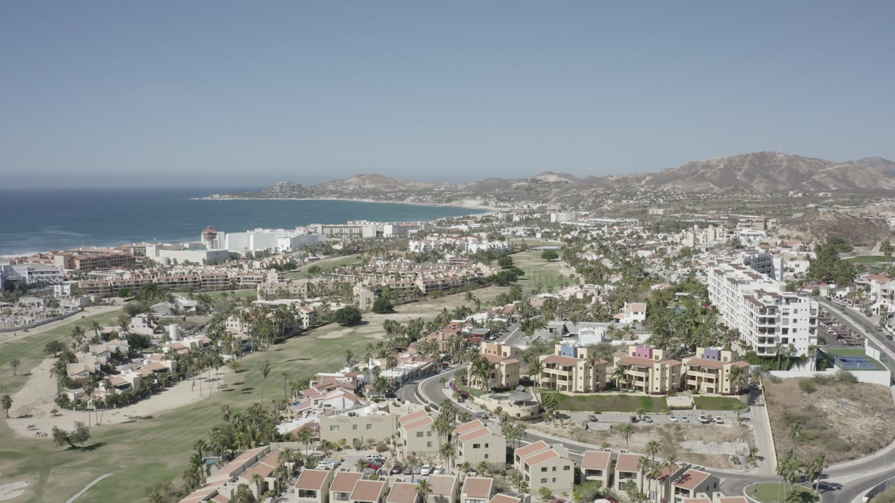 Opening aerial frame over San Jose del Cabo, Mexico, showing low-rise buildings, a white hotel complex, green landscaped areas, roads, dry hills, blue sea, and no visible people.