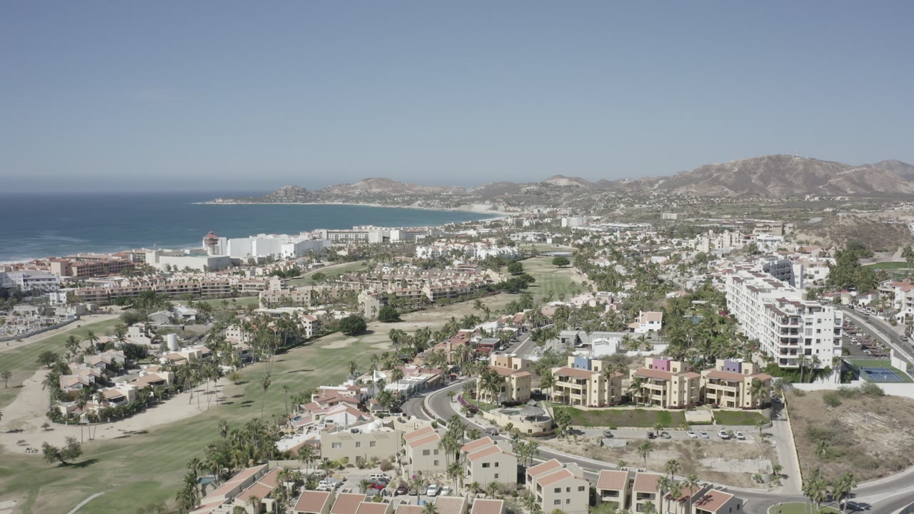 Wide drone view of coastal town, white buildings, roads, greenery, sea, and hills.