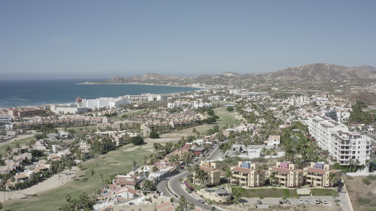 Aerial view of San Jose Del Cabo one second in, with the coastline held on the left, clustered white buildings across the center, a road and homes below, and no visible people.