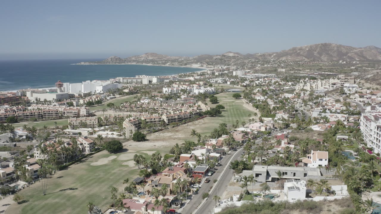 Opening aerial frame over San Jose del Cabo, Mexico, with homes and roads in the foreground, golf greens in the middle distance, a blue bay to the left, dry hills beyond, and no visible people.