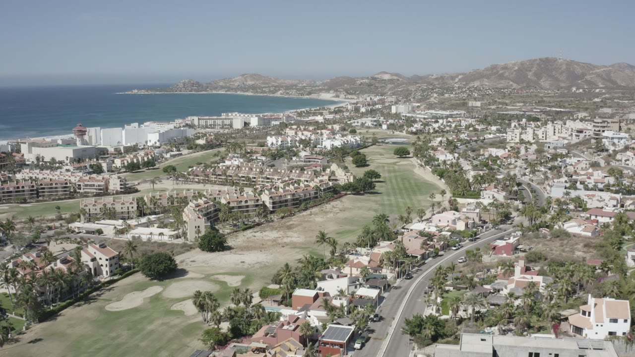 Aerial frame over San Jose del Cabo with the ocean on the left edge, white shoreline buildings, fairways through the center, residential rooftops below, and no visible people.