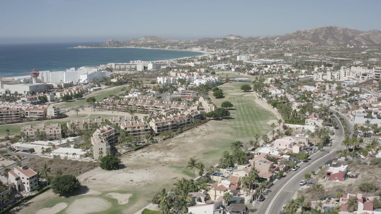 Wide aerial view over San Jose del Cabo with blue ocean at left, white beachside buildings, tan-roofed homes, golf fairways, curving roads, distant hills, and no visible people.
