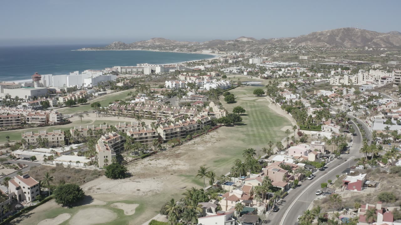 Aerial frame over San Jose del Cabo showing the ocean and sandy curve of coast behind white buildings, with residential blocks, golf course fairways, roads, hills, and no visible people.