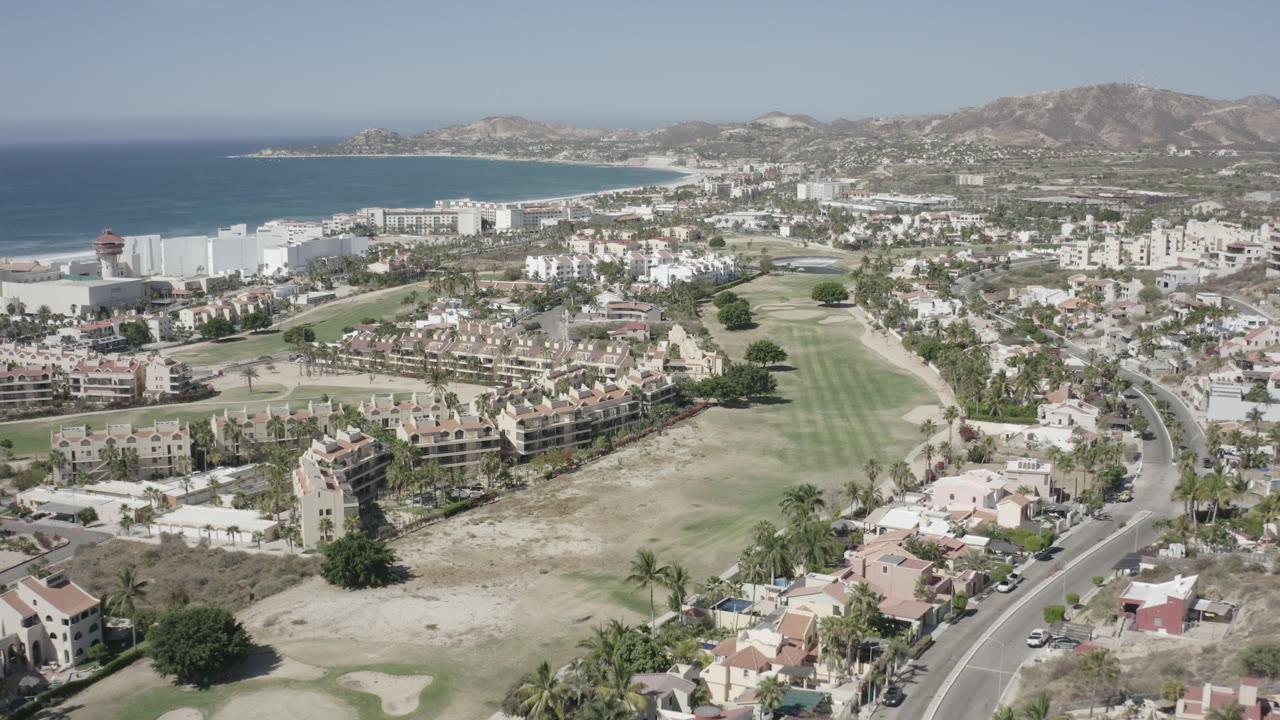 Drone-style view across resort buildings, homes, golf course, ocean, and hills.