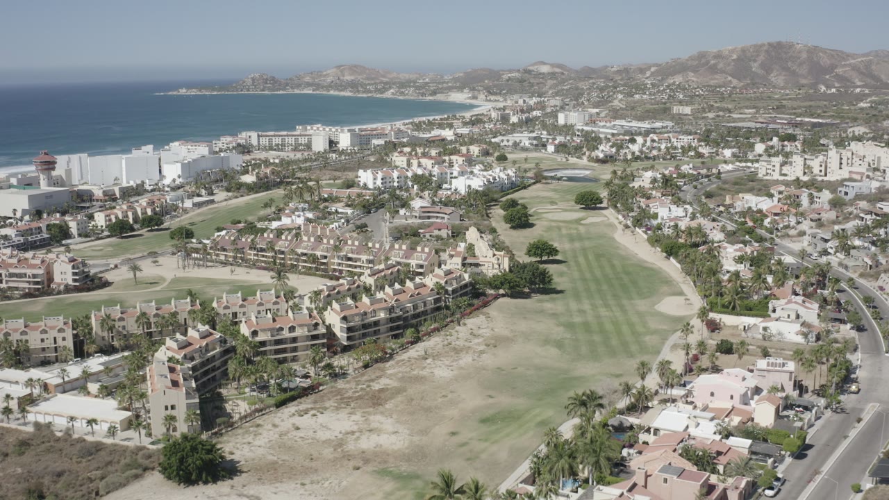 Wide aerial frame over San Jose del Cabo showing a golf fairway, apartment blocks, houses, ocean, and hills under clear daylight, with no visible people.