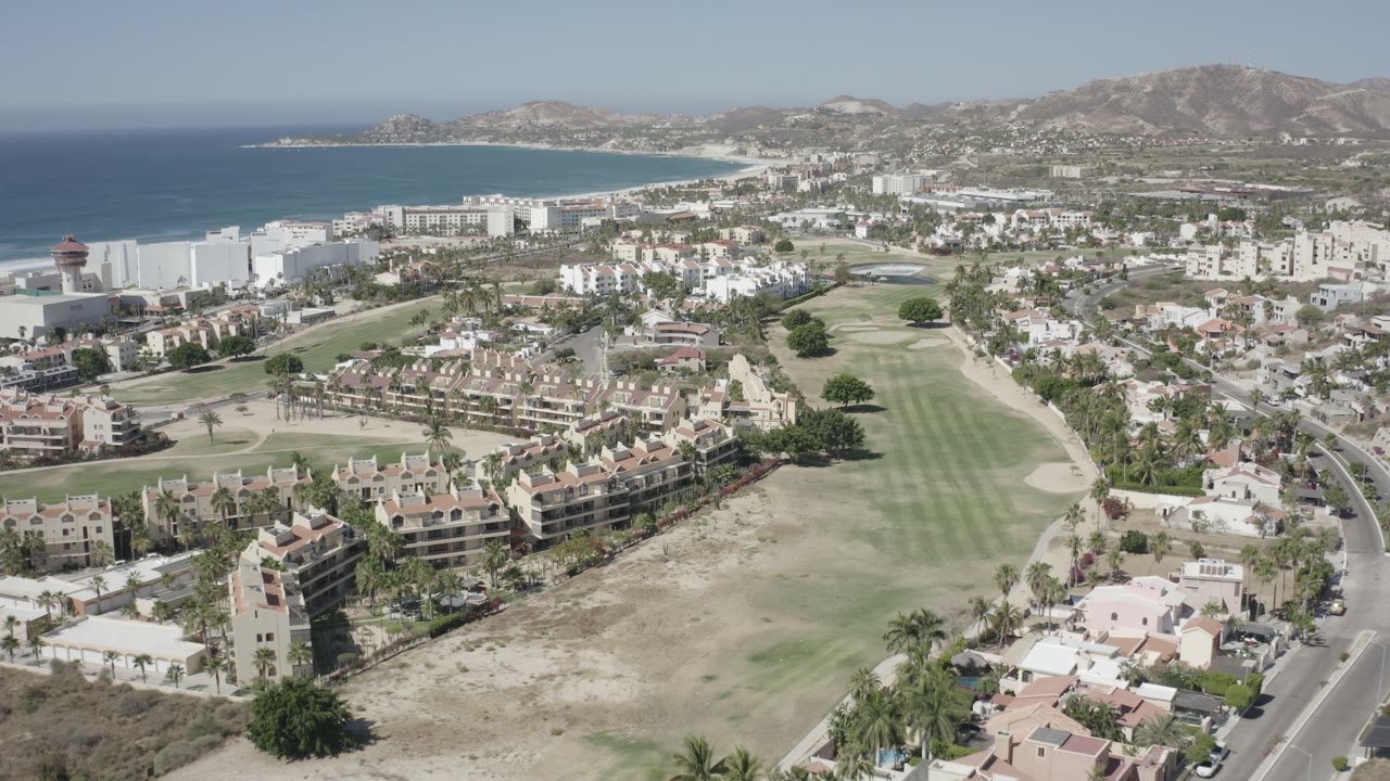 Aerial view in San Jose del Cabo with the fairway centered, residential roofs in the lower frame, large white coastal buildings to the left, and no visible people.